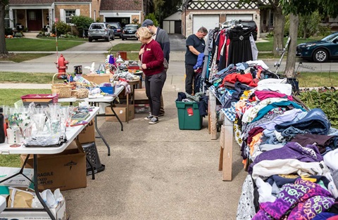 Residents browse through stacks of clothes on a table at a driveway garage sale in a residential neighborhood.