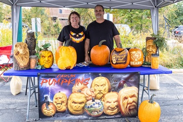 Two pumpkin carvers pose behind a table filled with carved pumpkins, showcasing their creations at the City of Des Plaines annual Harvest Hoot.