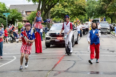 Patriotic street performers in the parade, including a stilt walker, unicyclist, and jugglers.