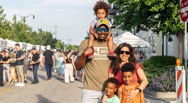 Image of a family with two parents and three children smiling at Taste of Des Plaines with food vendor tents in the background.